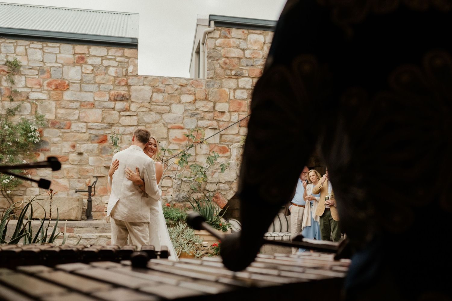 A candid wedding scene captured through a wooden deck railing with a stone wall backdrop.