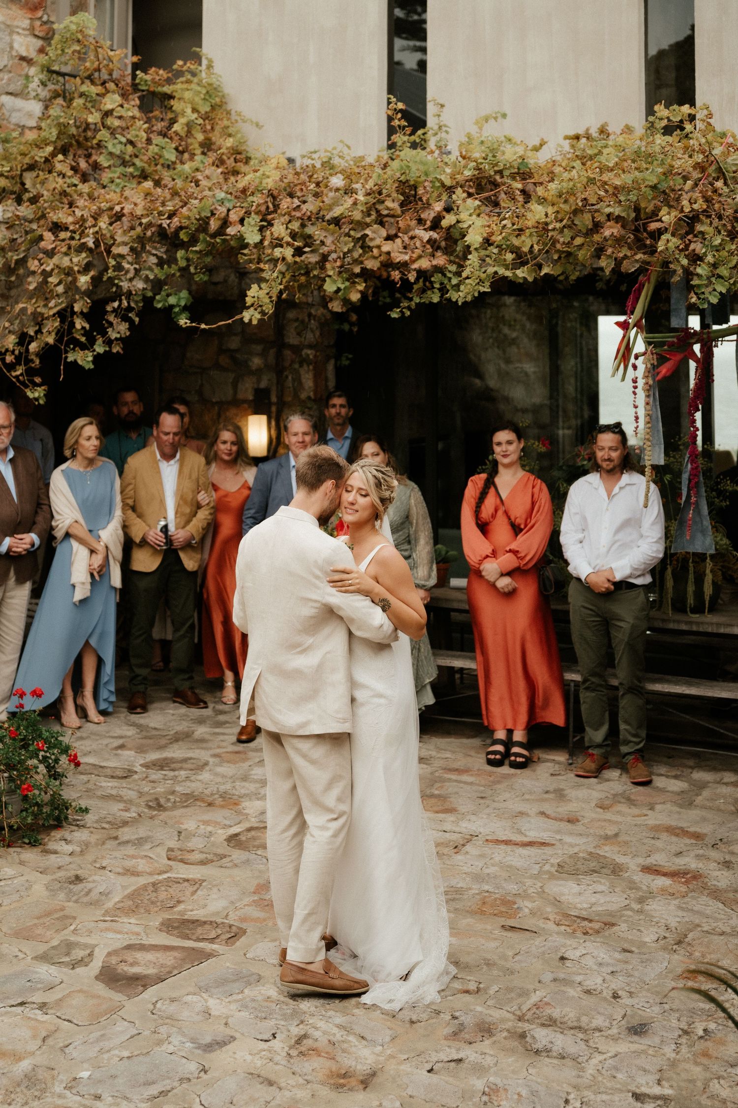 Couple shares their first dance surrounded by guests under floral decor at an outdoor reception.