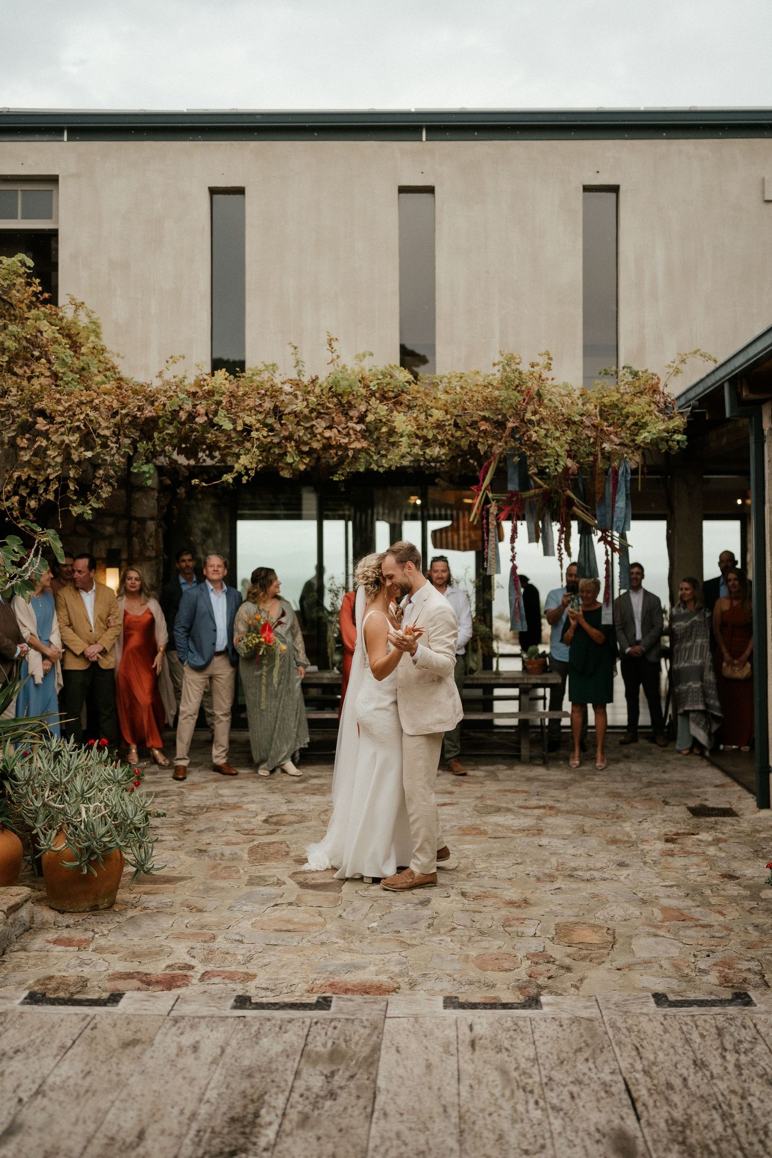 Newlyweds share their first dance on a stone patio surrounded by guests and lush landscaping.