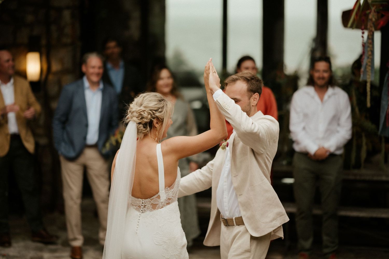 A wedding couple shares their first dance in a dimly lit indoor venue with guests watching in the background.