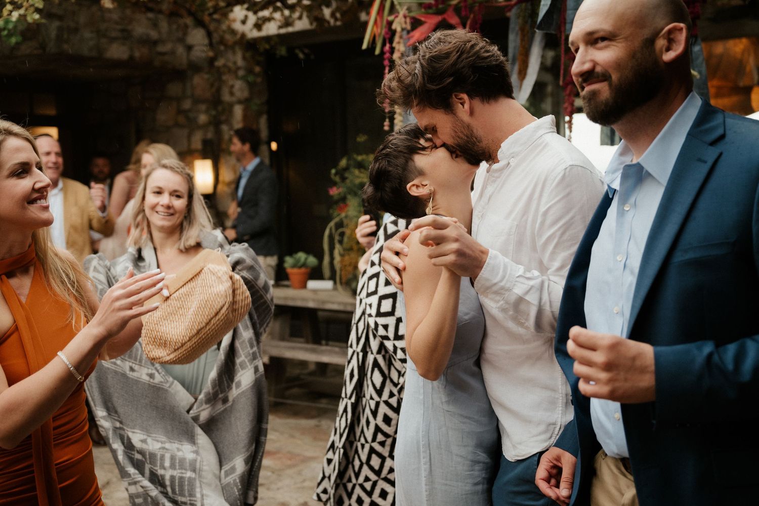 Wedding guests laugh and socialize at an outdoor reception with rustic stone walls.