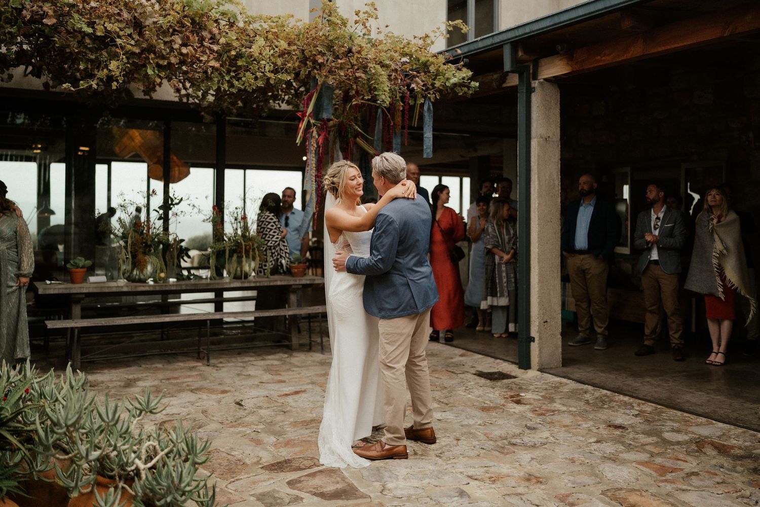 Couple dances together on outdoor patio with string lights and greenery in background.