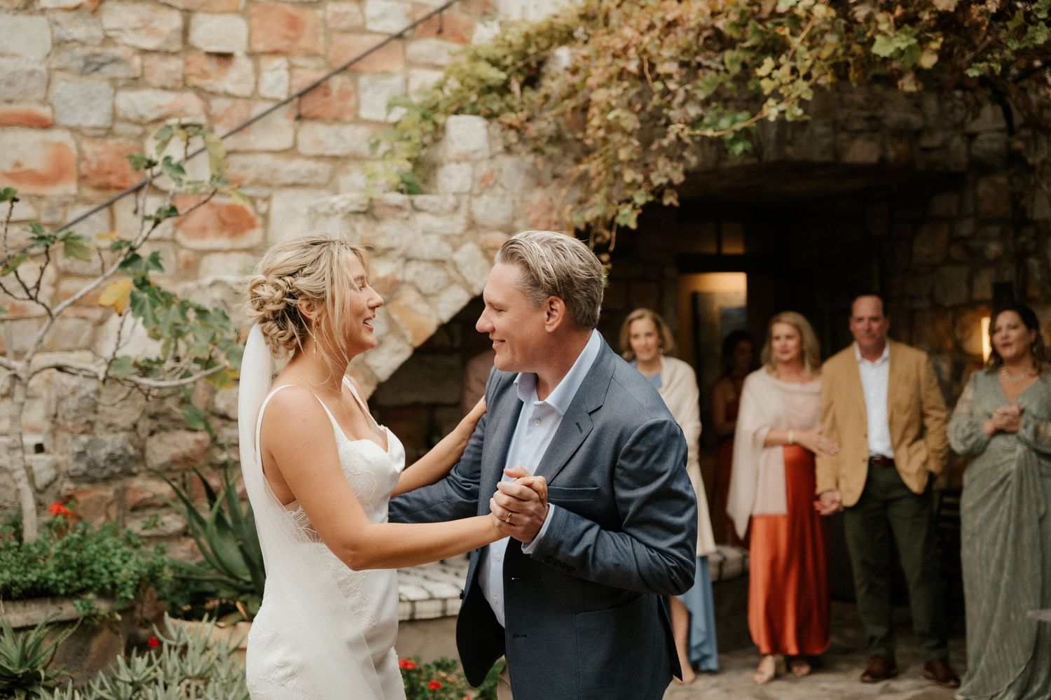 Couple sharing a dance moment in front of a rustic stone wall with guests watching.