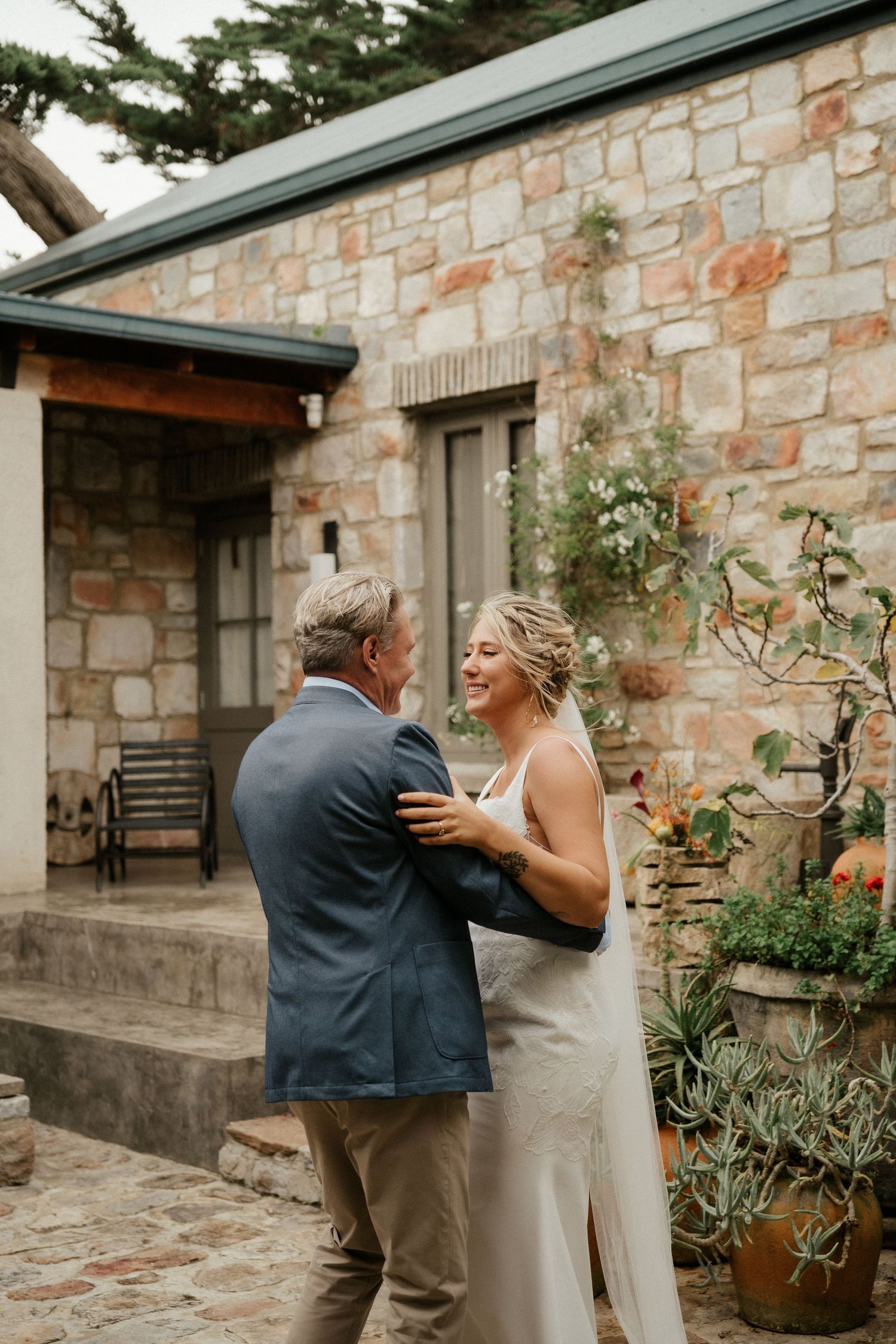 Two people share a joyful moment together on a stone patio outside a rustic stone building with climbing vines.