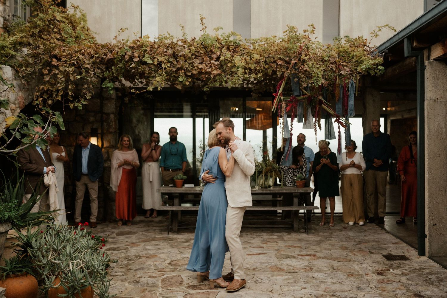 Couple dancing at an outdoor wedding reception under a vine-covered pergola.