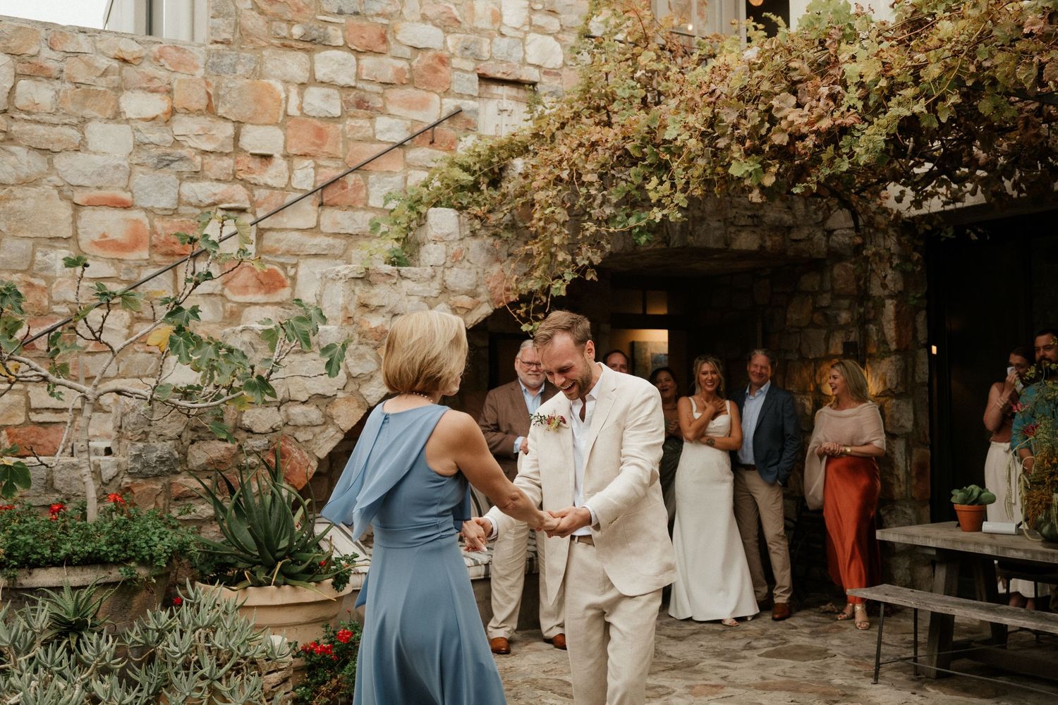 Wedding guests mingle outside a rustic stone building with climbing vines and flowers.