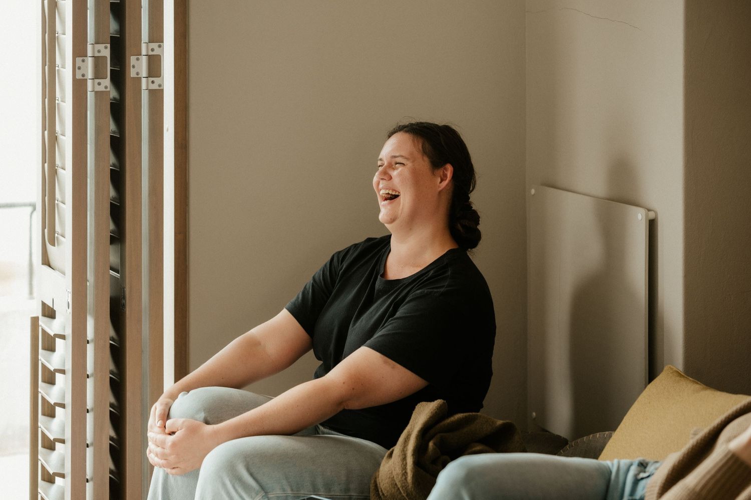 Someone sits by a window laughing joyfully while relaxing in casual attire.