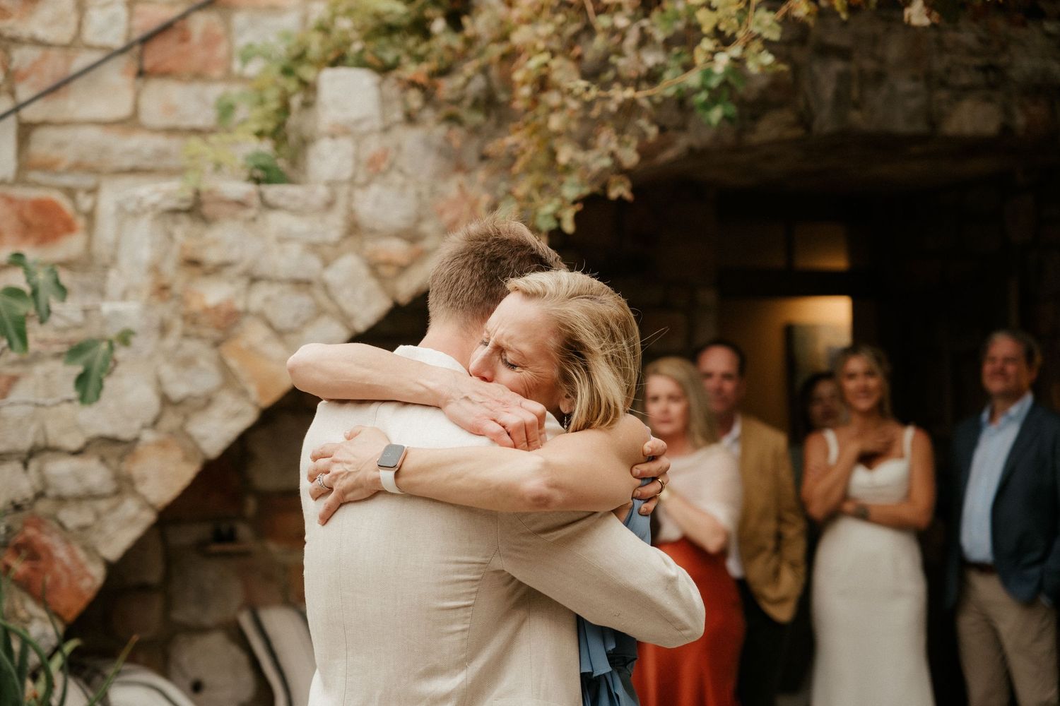 Emotional embrace at wedding celebration with stone wall backdrop and onlooking guests.