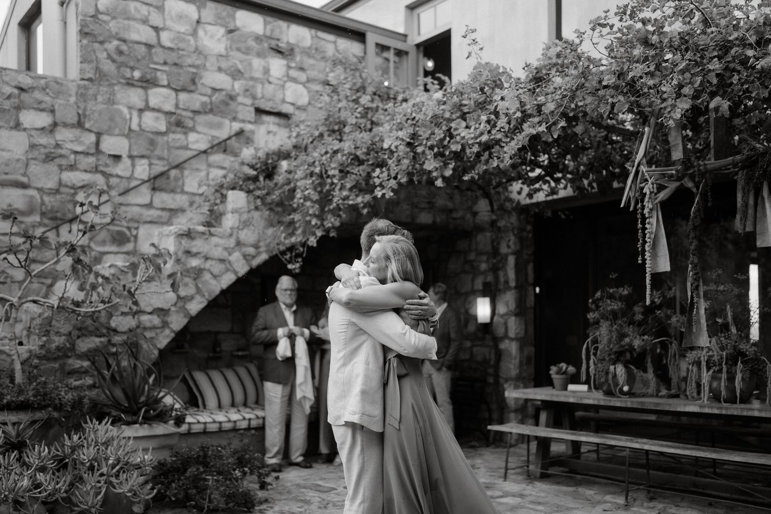 A romantic embrace captured in black and white against a rustic stone wall backdrop.
