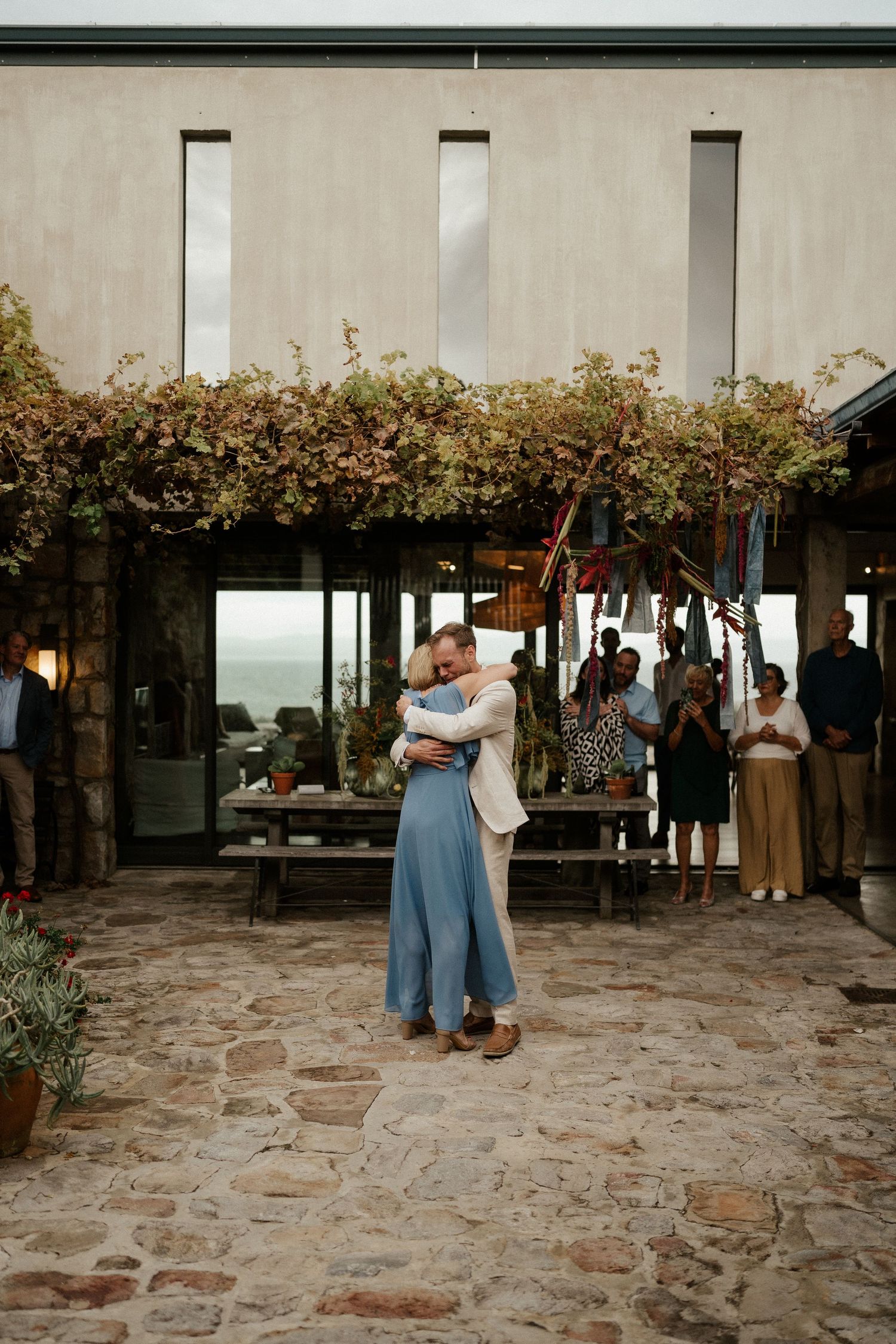 Couple embracing on rustic stone patio with greenery-covered walls and guests mingling in background at outdoor venue.