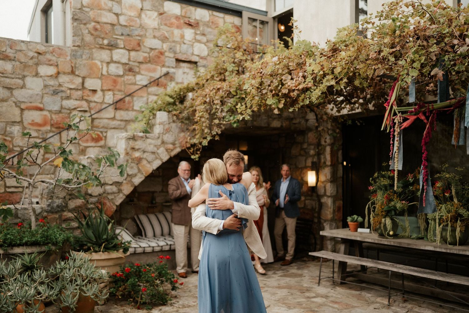 Romantic moment between couple during outdoor wedding reception at stone-walled venue with climbing vines.