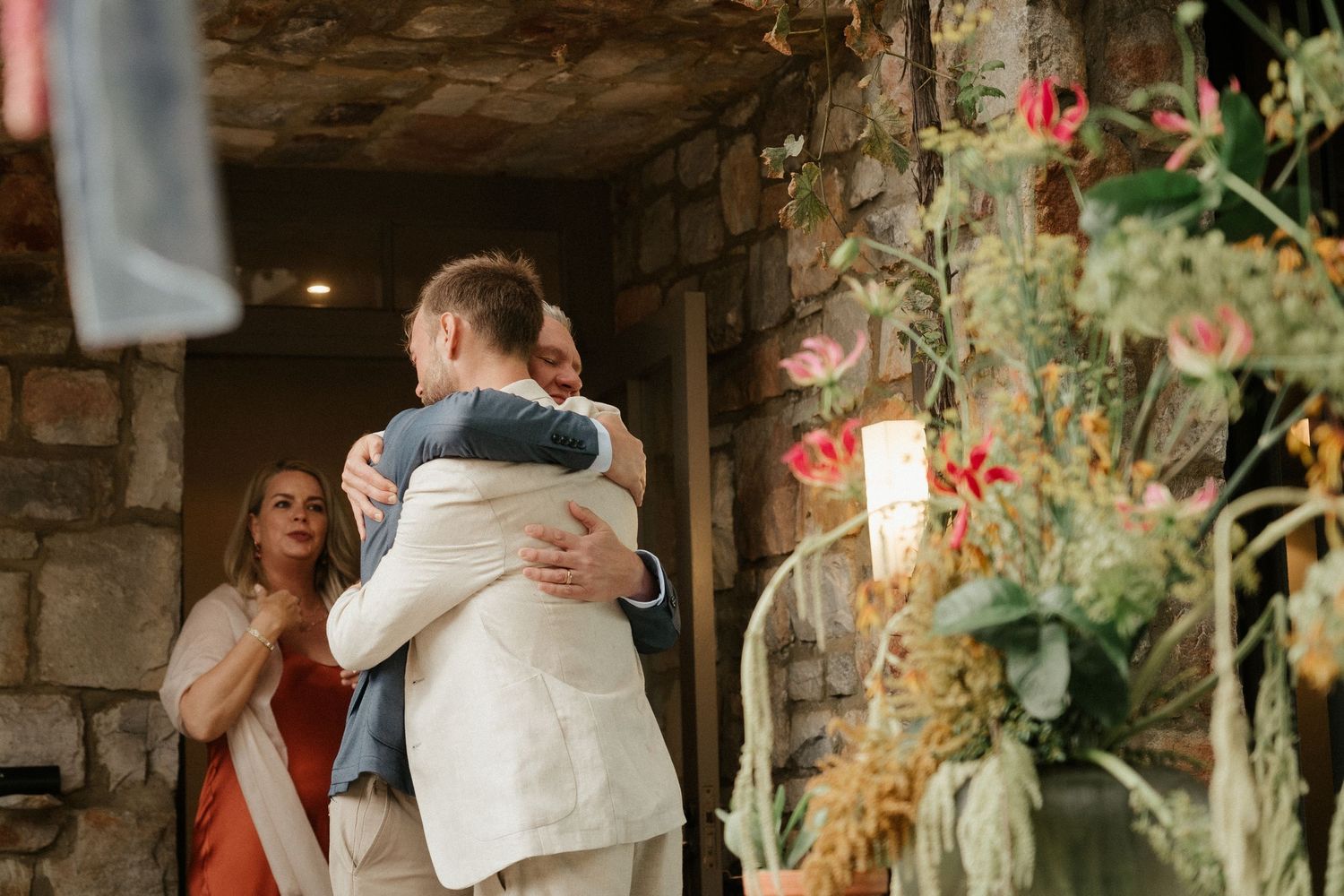 Two people embrace in an emotional hug at a wedding ceremony surrounded by floral decorations and candlelight.