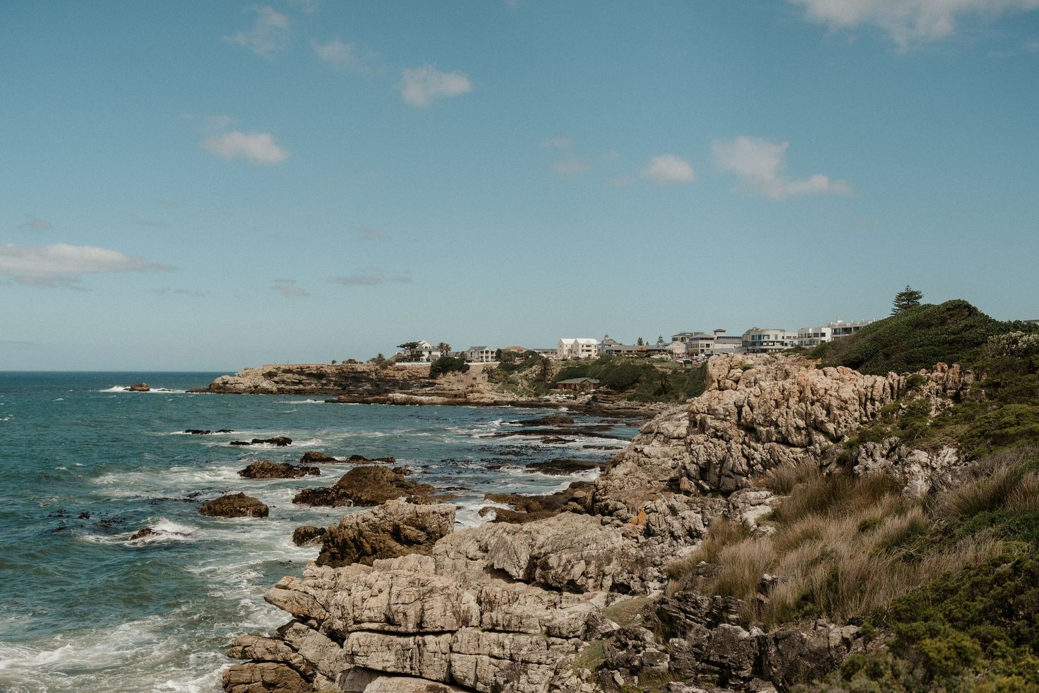 Rocky coastline with turquoise waters and white buildings perched on cliffs under blue sky with scattered clouds.