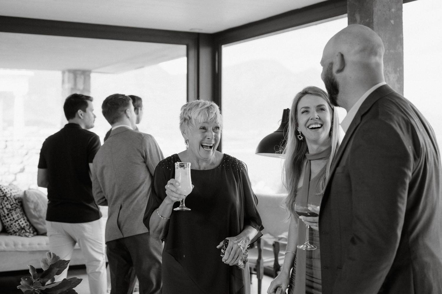 A candid black and white photo captures guests laughing during a joyful wedding reception moment.