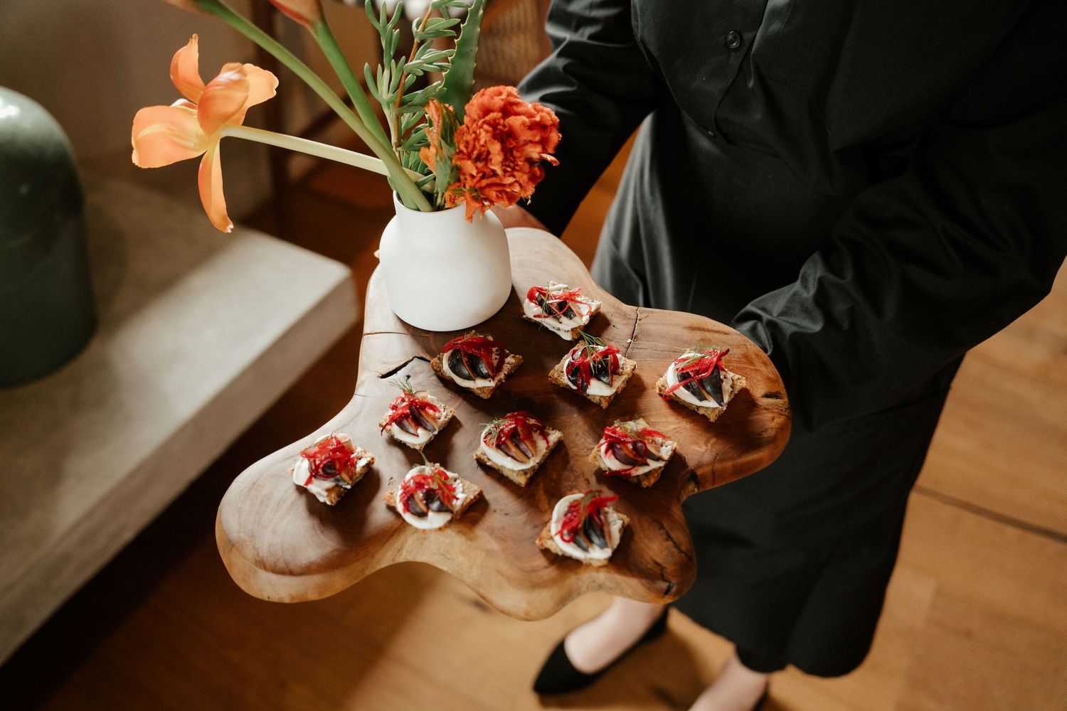 Elegant hors d'oeuvres displayed on wooden serving board with orange flowers in white vase.