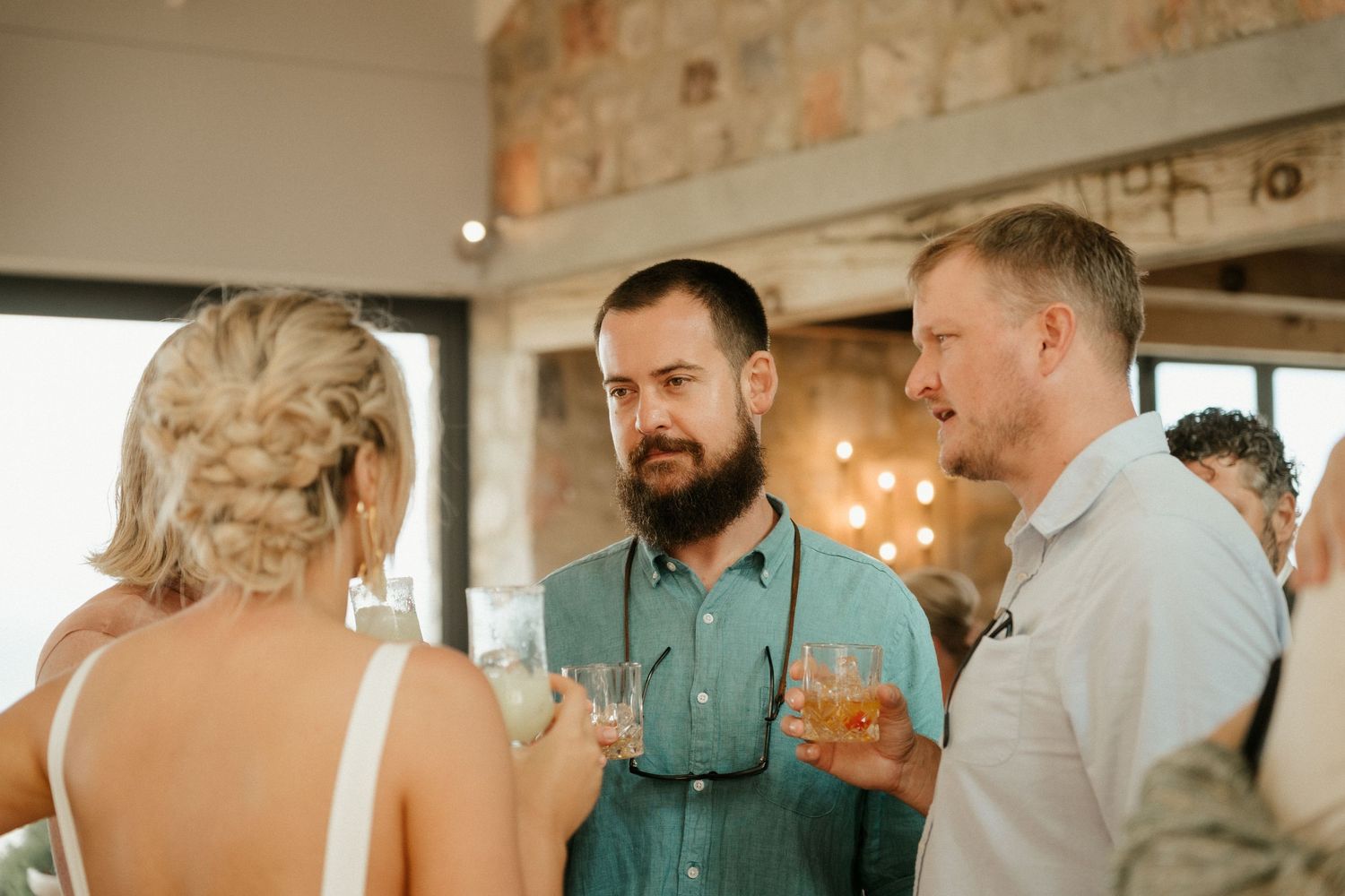 Guests chatting and holding drinks at an indoor wedding reception with stone walls.