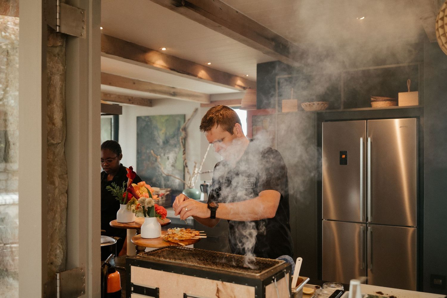 Chef cooking at indoor event with smoke rising from grill in modern kitchen setting.