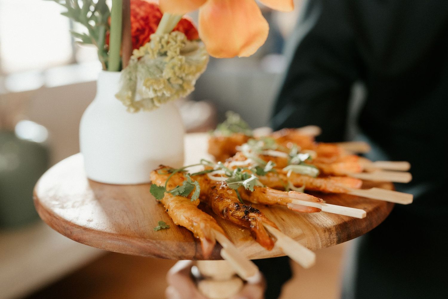 A wooden serving board displays a neatly arranged appetizer spread with orange flowers in a white vase.