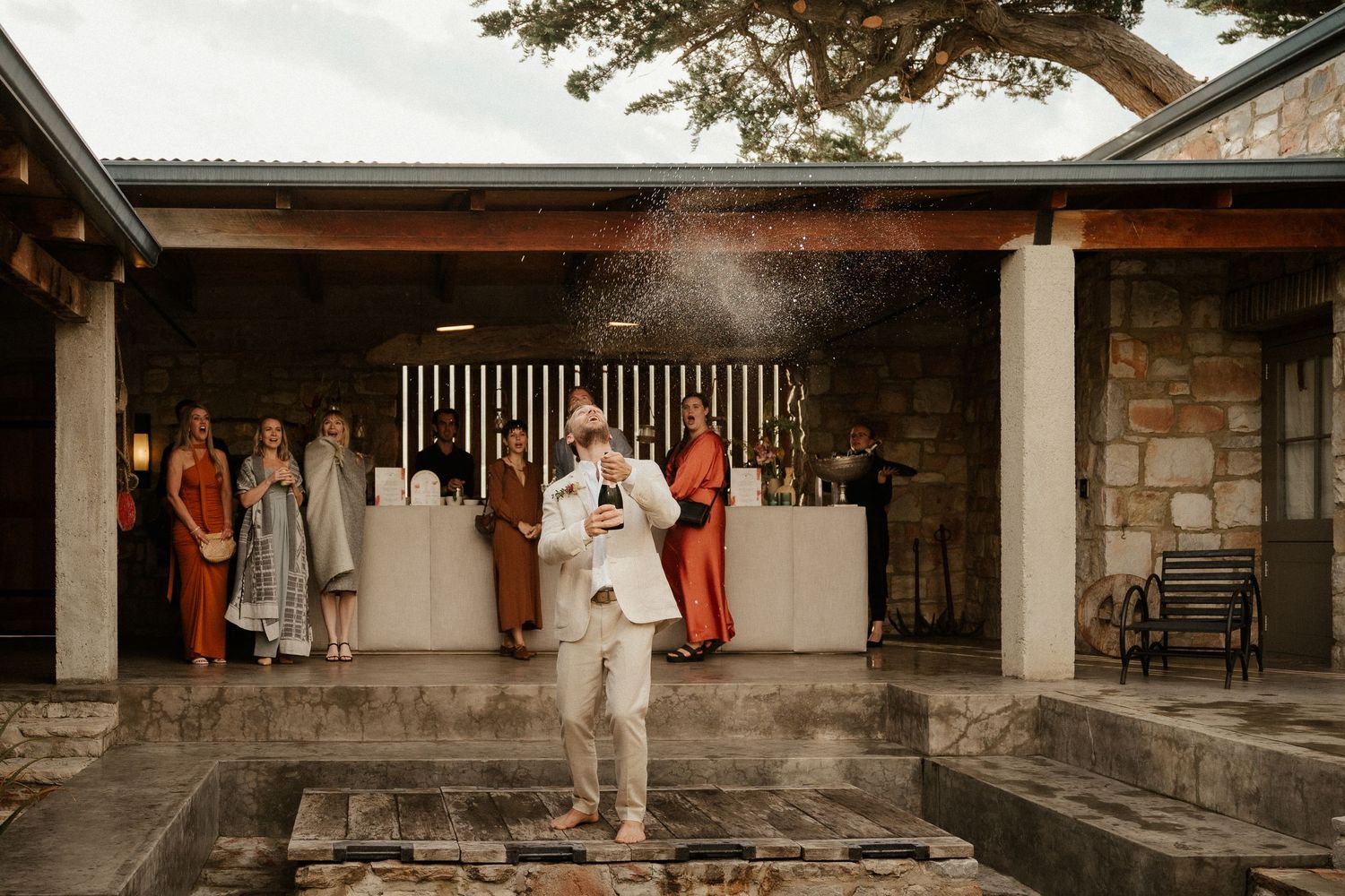 A person in white celebrates on a stone patio with wedding guests and decor in the background.