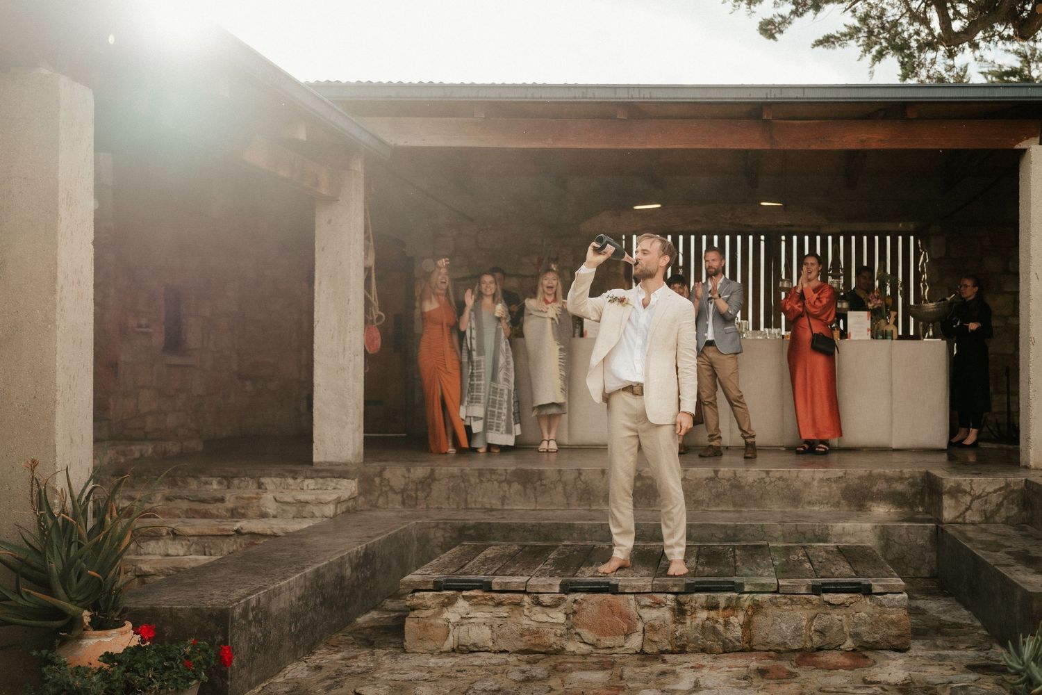 Wedding guest in all-white suit poses on stone steps with guests gathered behind in warm sunset light.