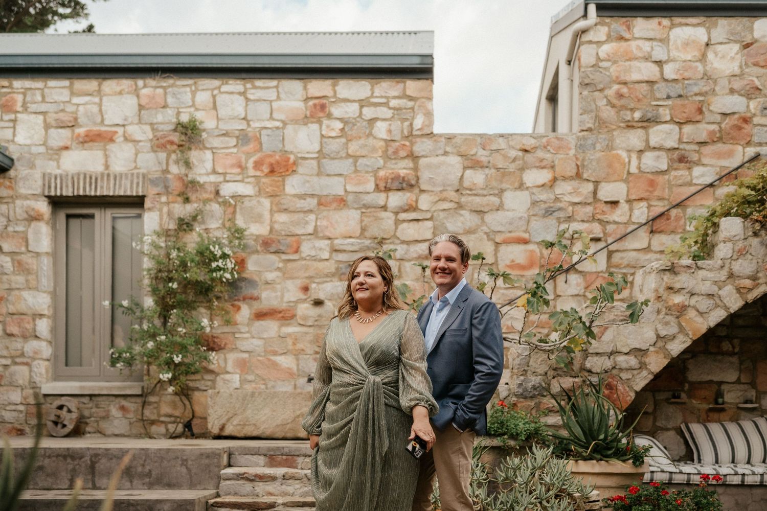 A couple poses against stone wall with climbing vines at elegant venue.