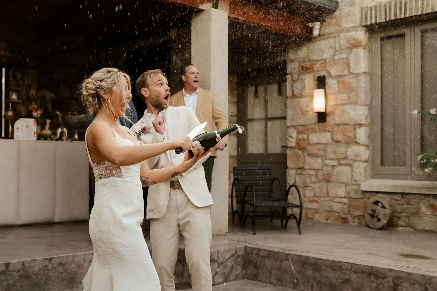 Wedding celebration with guests popping champagne bottles on a stone patio.