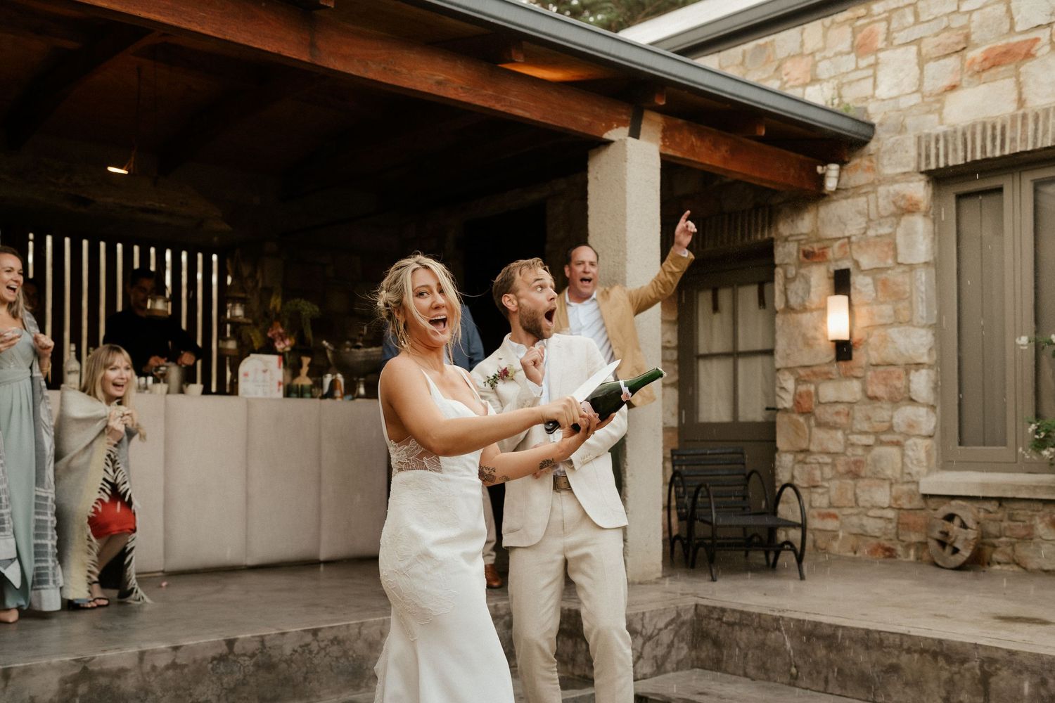 Couple popping champagne during outdoor wedding celebration at rustic venue.