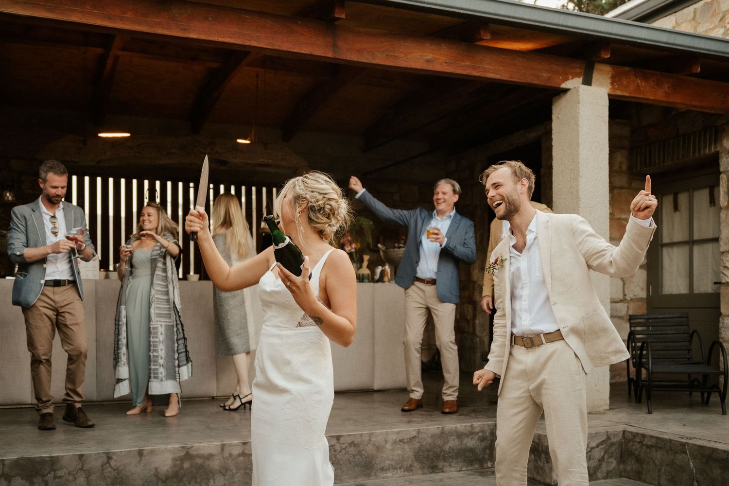 A couple shares a joyful first dance at their reception while guests look on under a rustic covered patio.