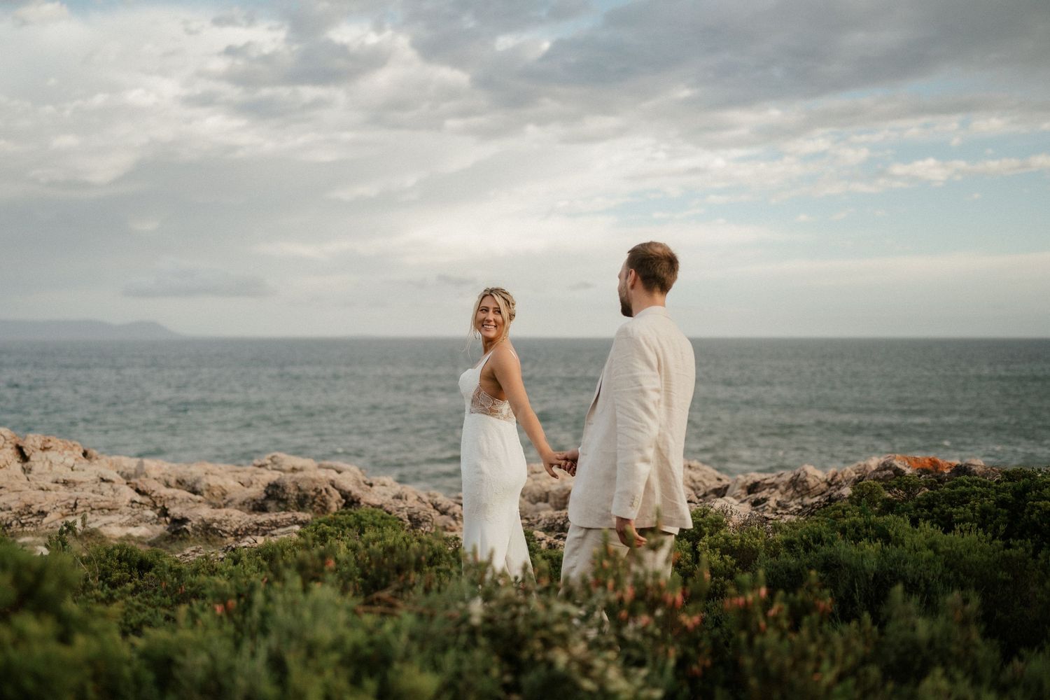 A couple in wedding attire stands together overlooking ocean coastline.