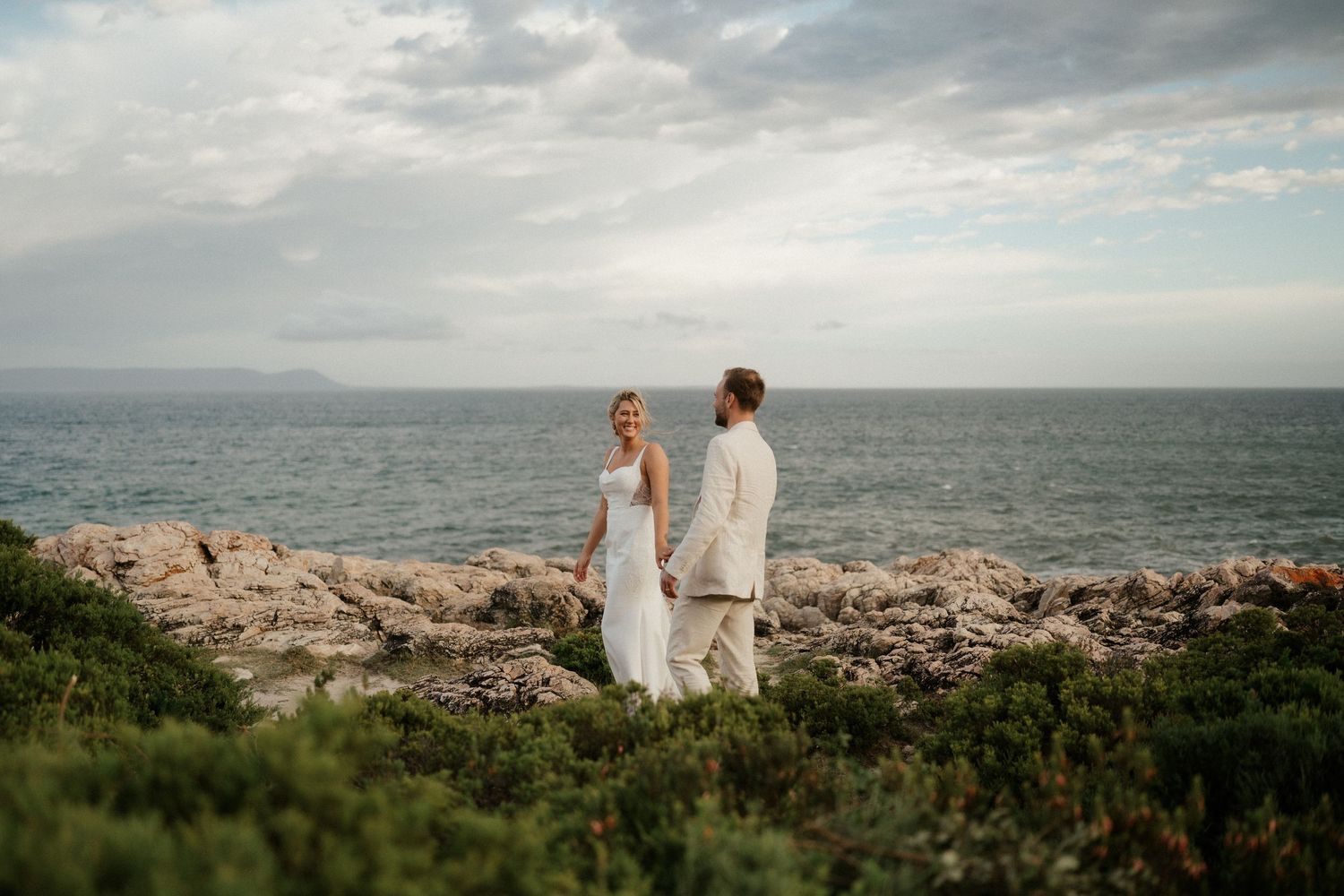 Coastal wedding portrait with couple standing on rocky cliff overlooking ocean at sunset.