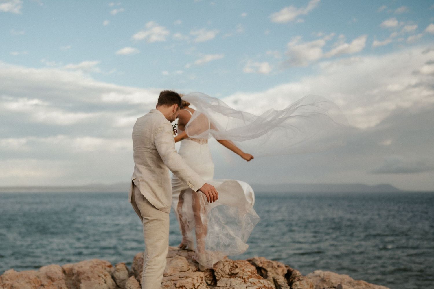 A romantic wedding photo session on coastal rocks with flowing veil and ocean backdrop.