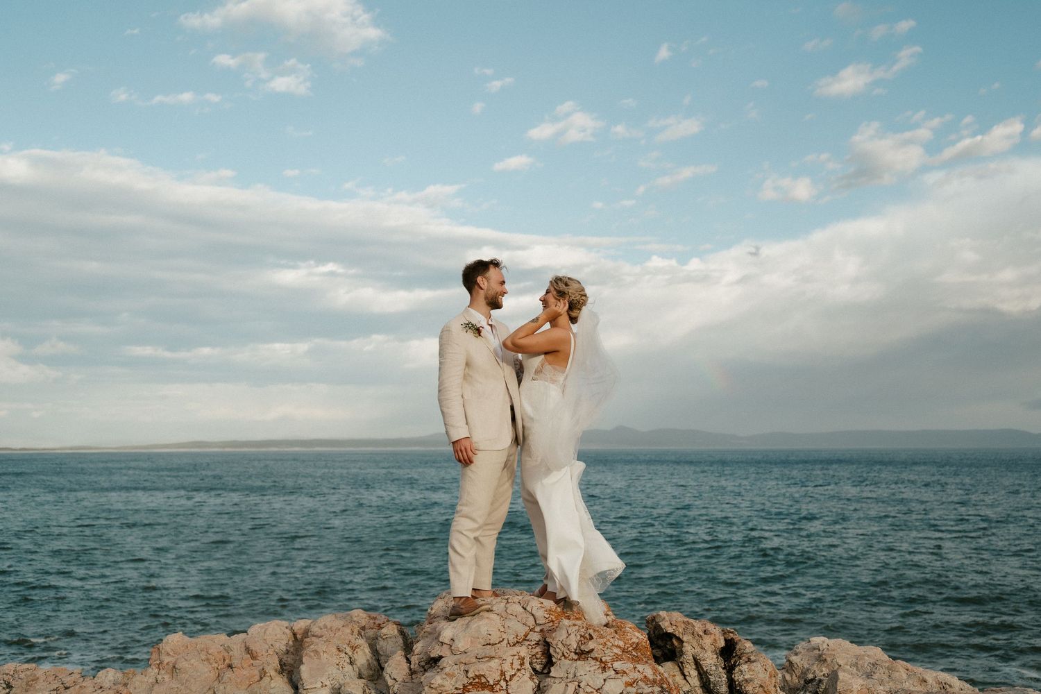 Wedding couple stands on rocky coastal outcrop against blue sky and ocean backdrop.