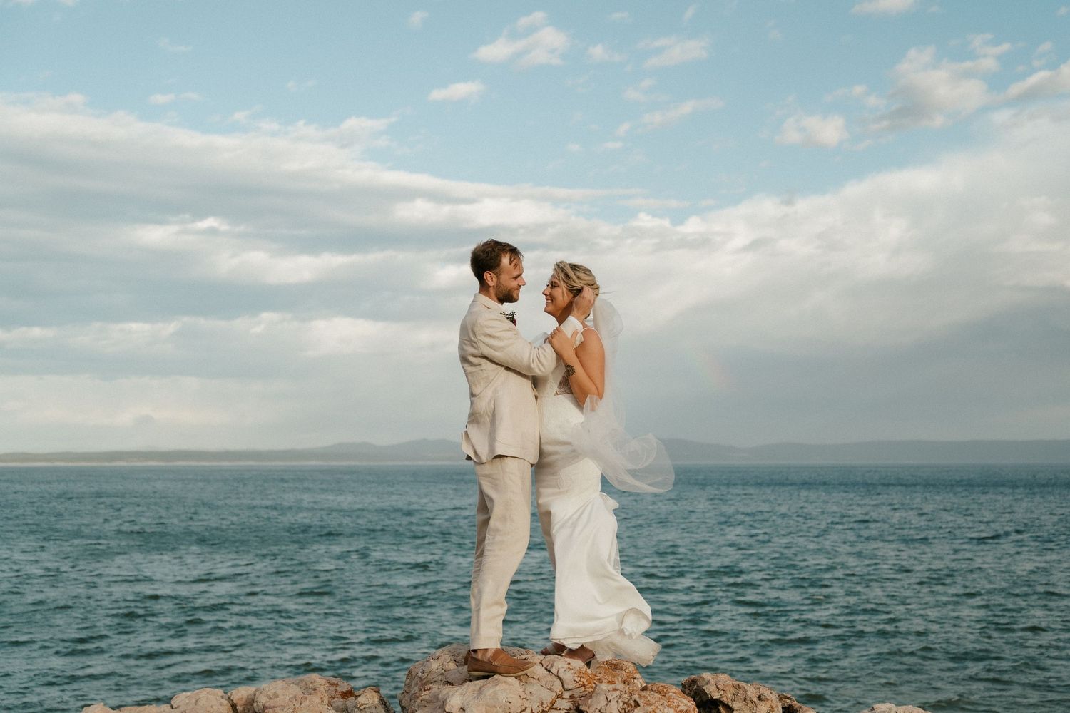 Couple standing on coastal rocks with ocean and cloudy sky backdrop at sunset.