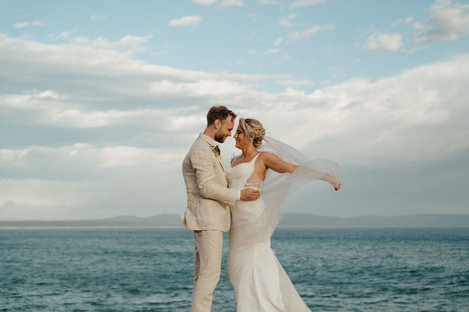 Couple in light colored wedding attire share an intimate moment against an ocean backdrop.