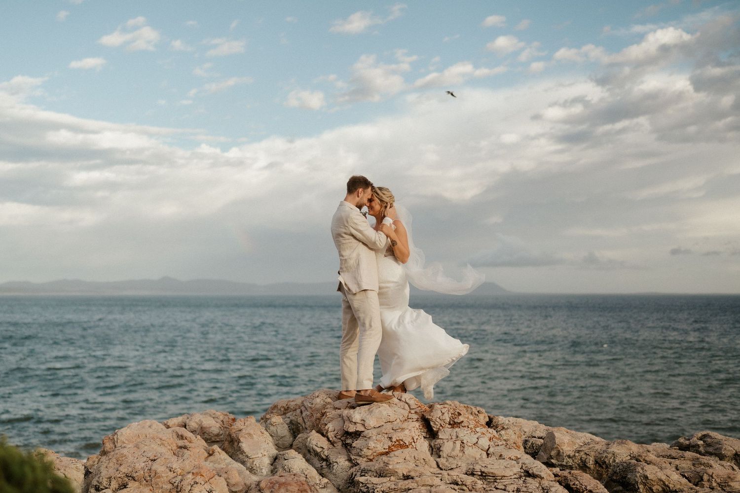 Couple embraces on rocky oceanfront during their coastal wedding ceremony.