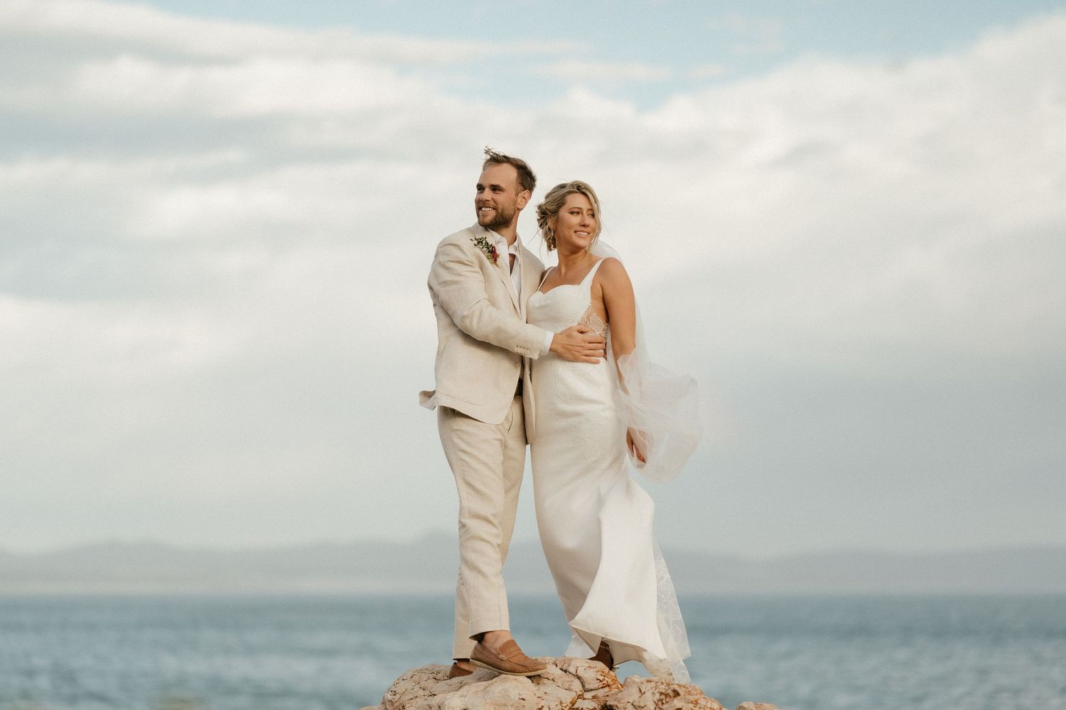 Couple in light colored attire pose on rocky oceanfront during cloudy day.