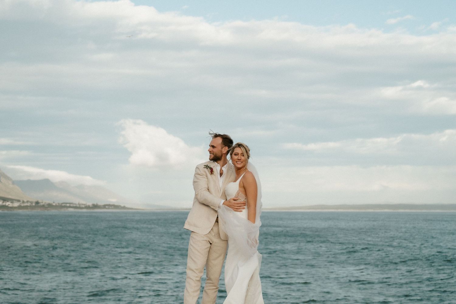 A romantic wedding photo of a couple embracing by the ocean with mountains in the background.