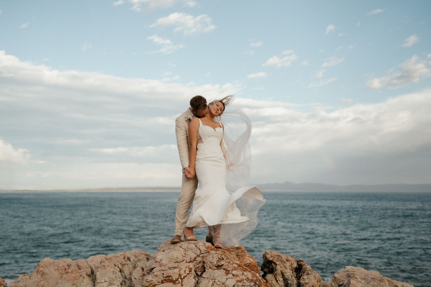 A couple shares a romantic moment on coastal rocks overlooking the ocean at sunset.