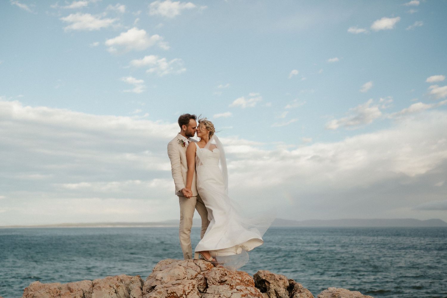 A couple in wedding attire stands on a rocky cliff overlooking the ocean.