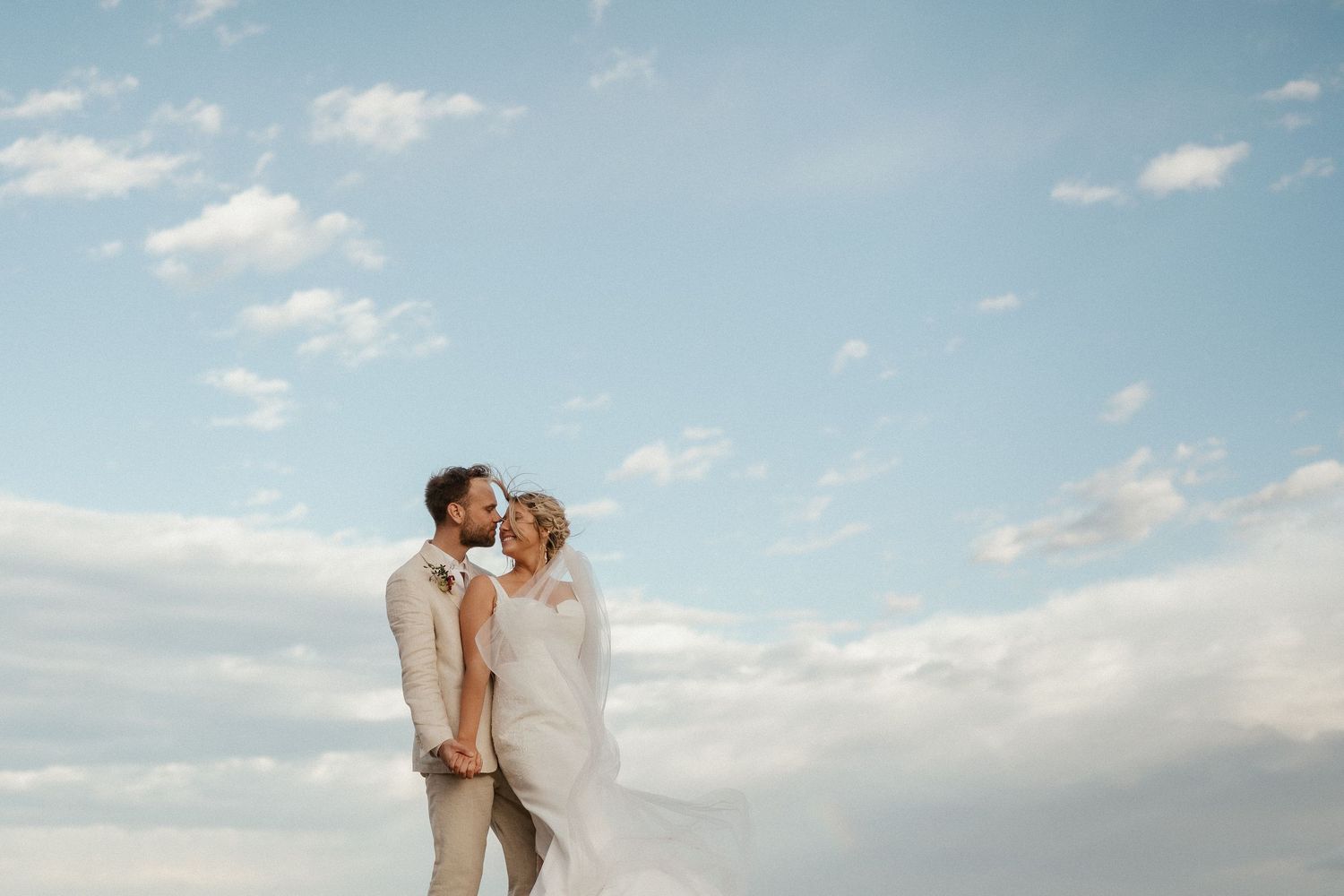 A couple shares a romantic moment against a bright blue sky with wispy clouds during their outdoor wedding.