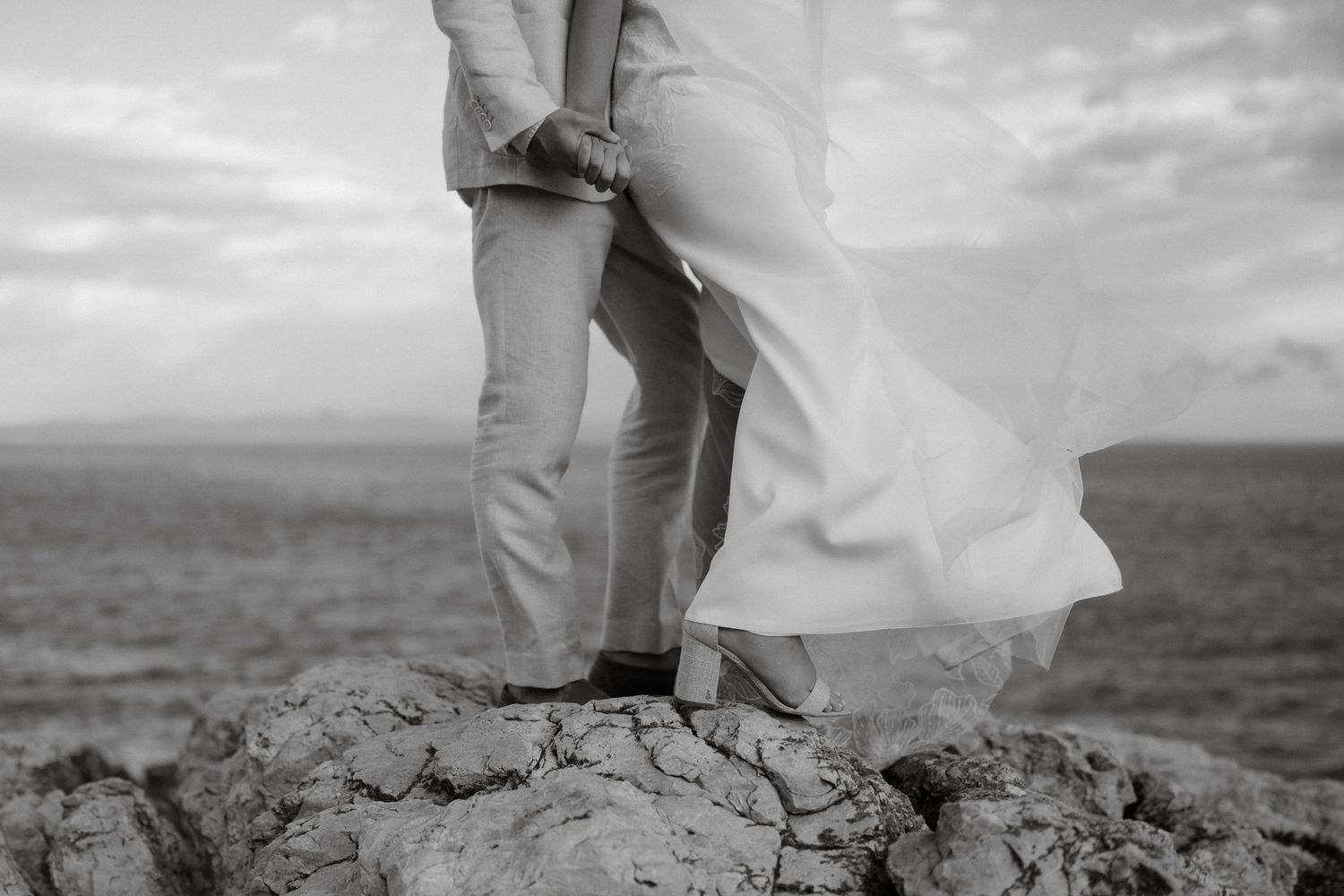Black and white image capturing flowing dress fabric against rocky coastal landscape.