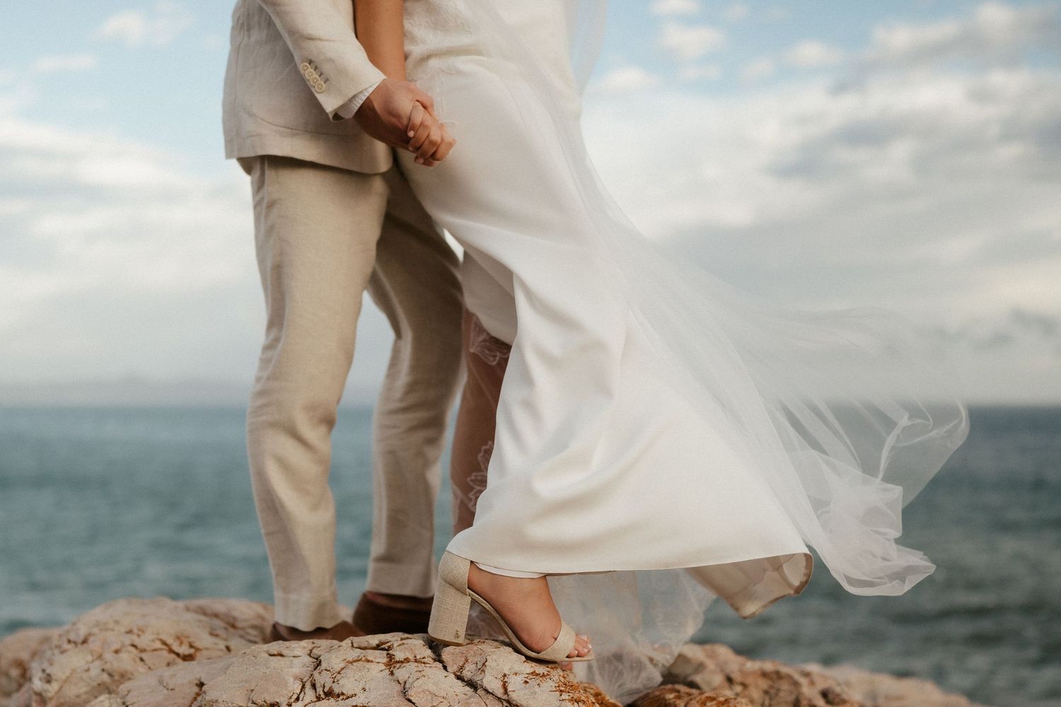 A romantic detail shot of wedding attire and flowing dress fabric against an oceanside cliff.