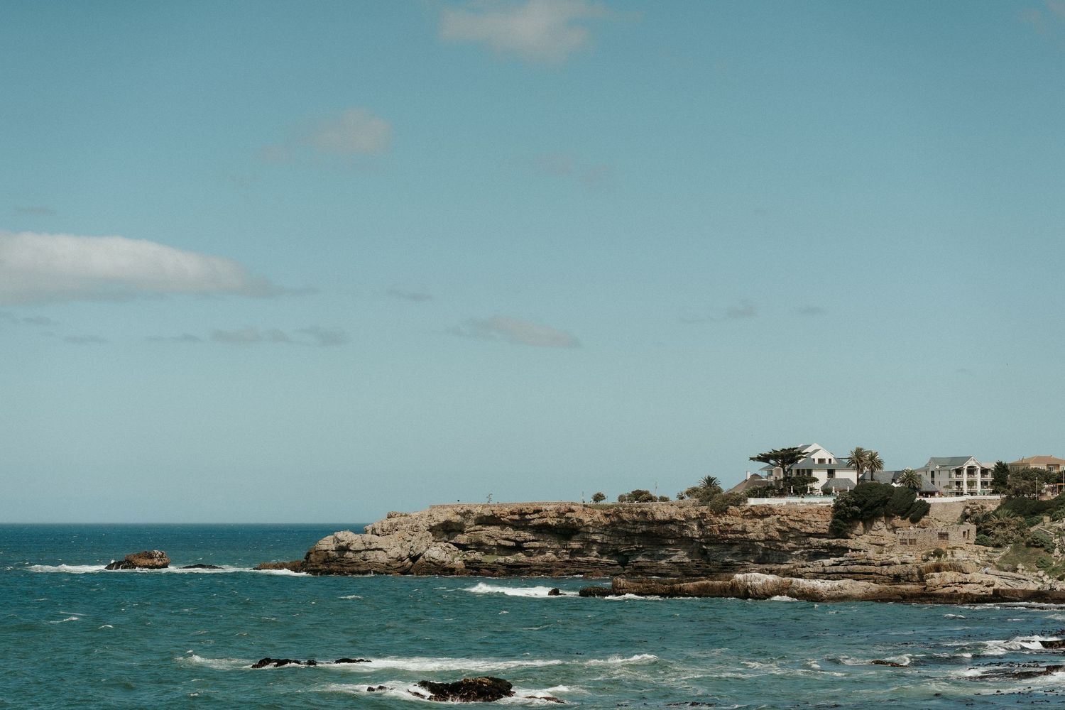 Scenic coastal landscape with rocky cliffs, ocean waves, and houses perched along the shoreline.