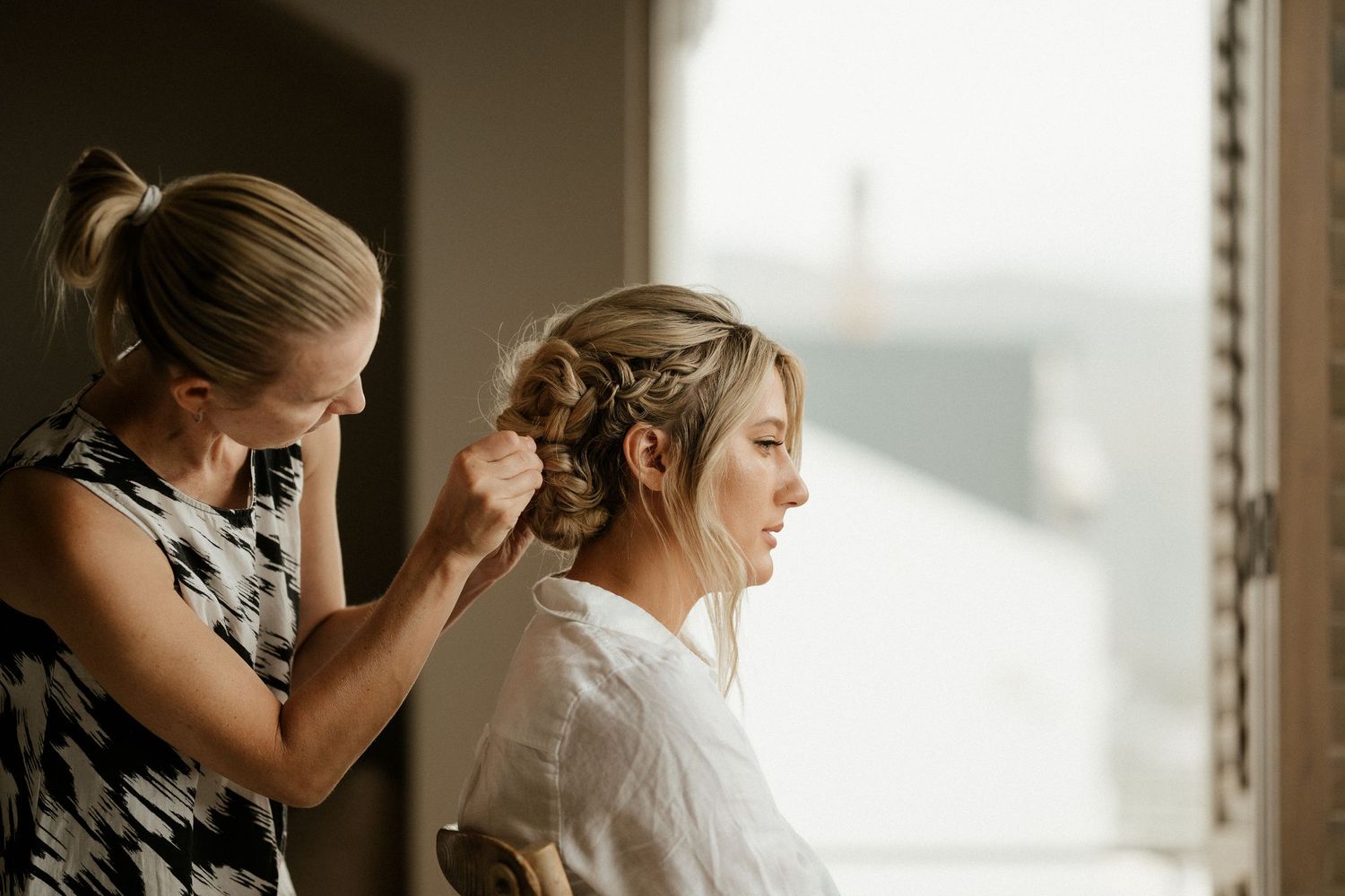 Wedding hairstylist works on bride's updo hairstyle by window in natural morning light.