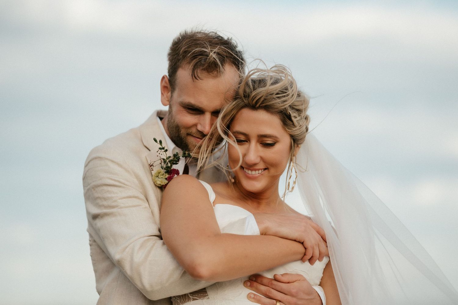 Close-up of a tender embrace between newlyweds against a cloudy sky.