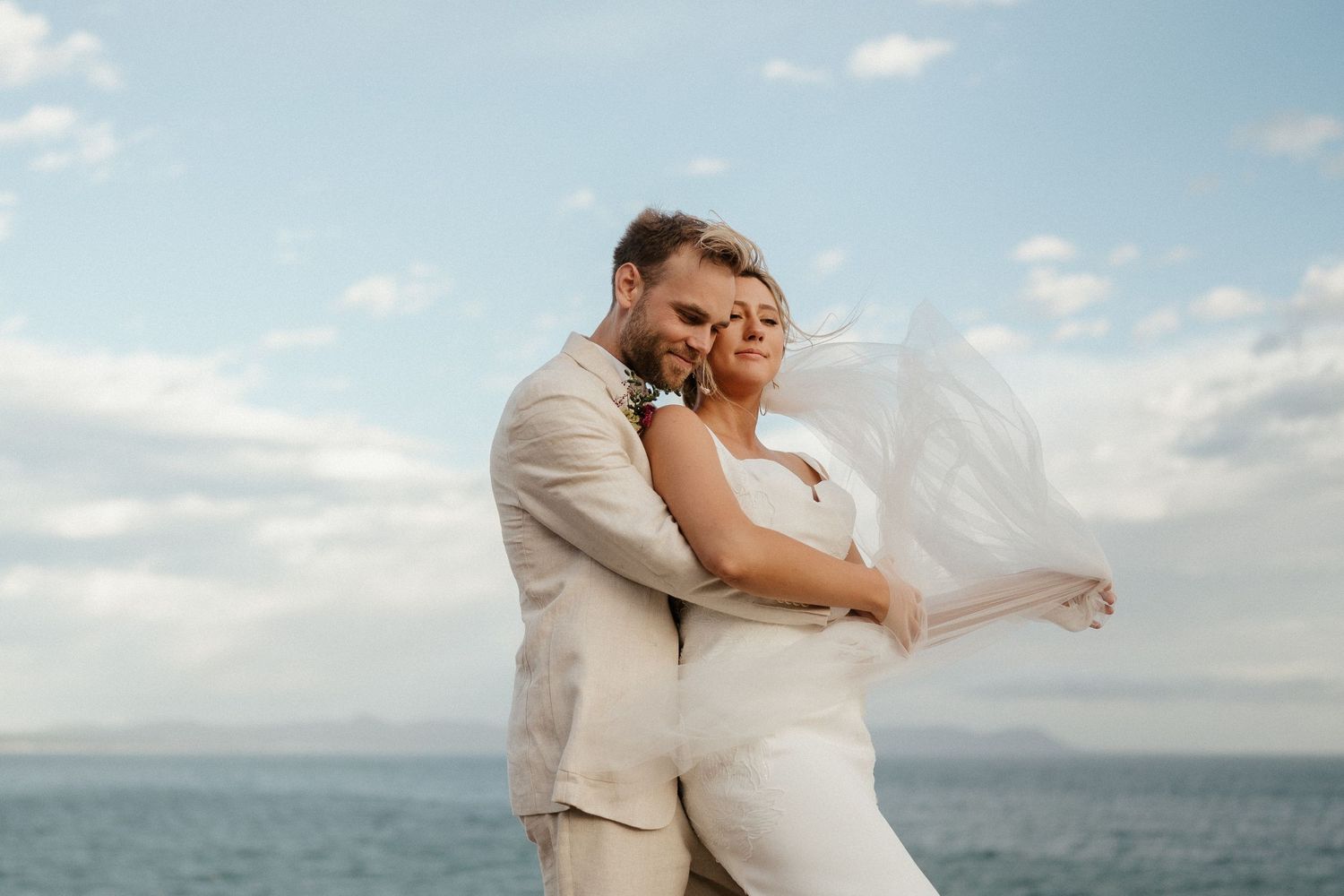 Romantic seaside moment with bridal veil flowing in the wind against blue skies.