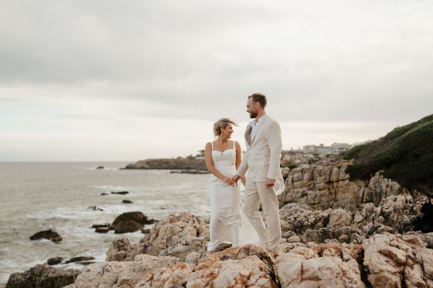 A couple stands together on rocky coastal cliffs overlooking the ocean on a cloudy day.