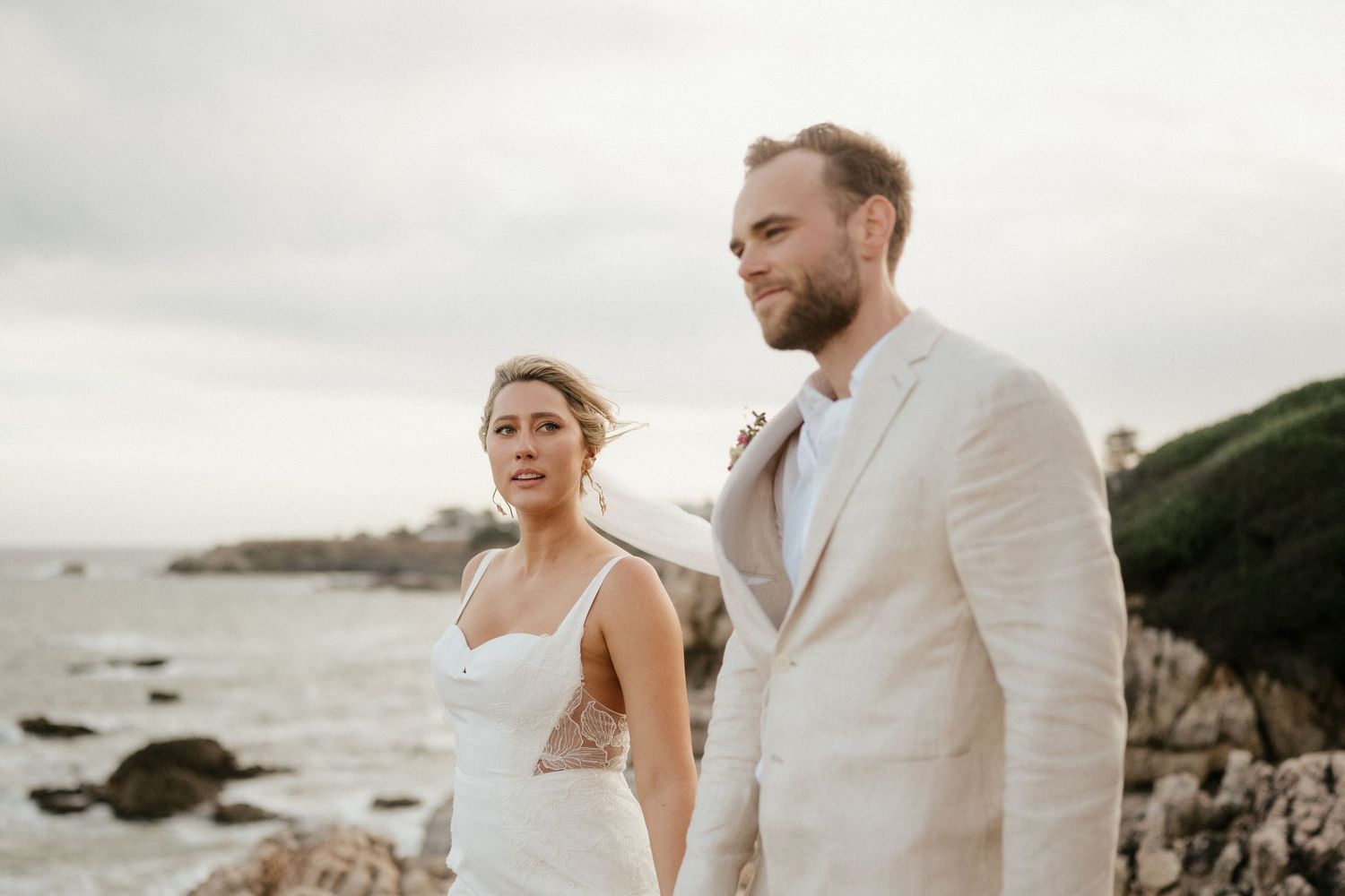 Couple in wedding attire standing on rocky coastline during overcast day.