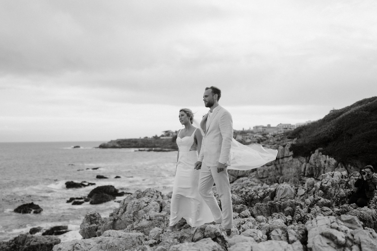 Couple in light-colored attire stands on rocky coastal cliff overlooking the ocean.