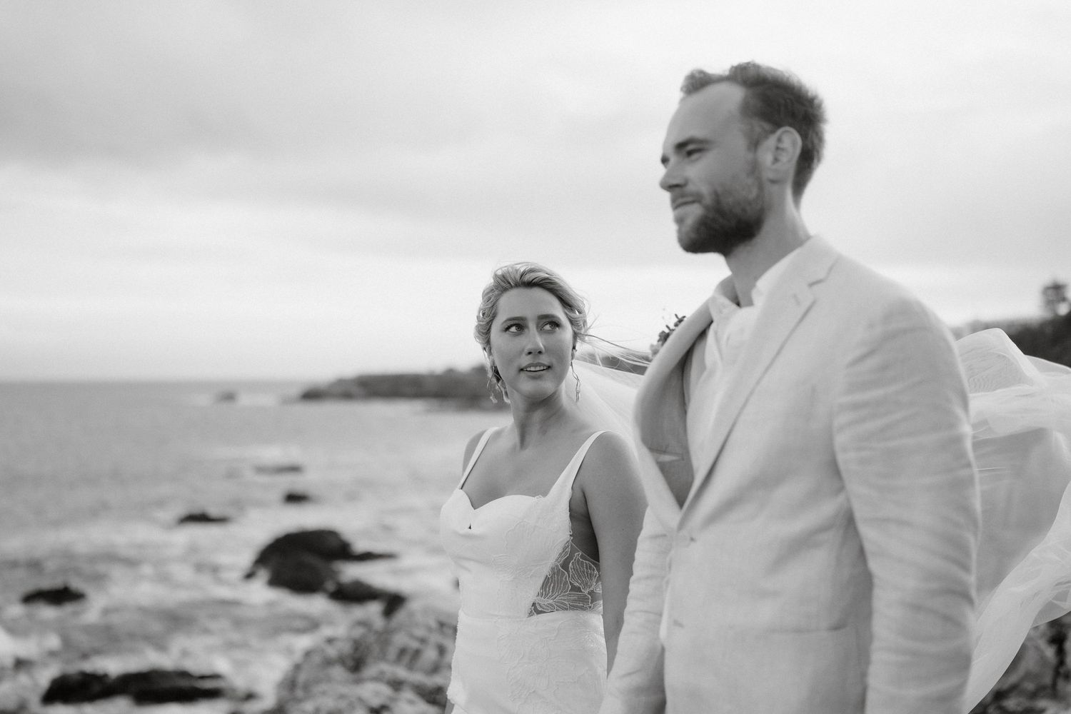 A couple in wedding attire standing near rocky coastline under cloudy skies.